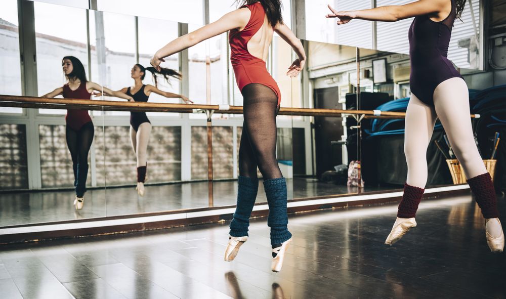 Crop women in bodysuits jumping on pointe above floor in studio training ballet.