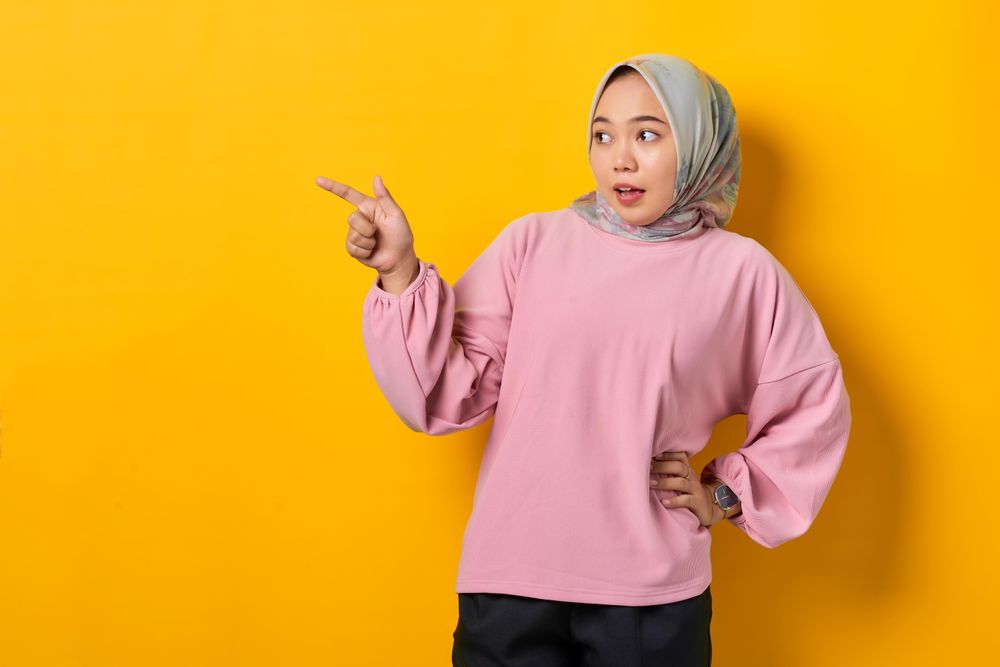 Surprised Muslim woman in headscarf pointing to space beside her with hand on hip