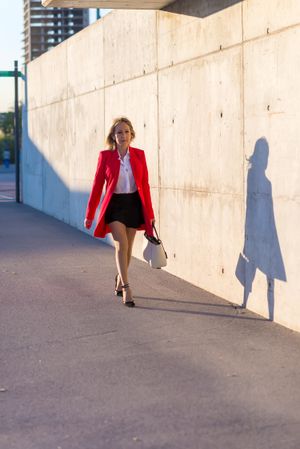 Businesswoman wearing red jacket walking next to cement wall with shadow
