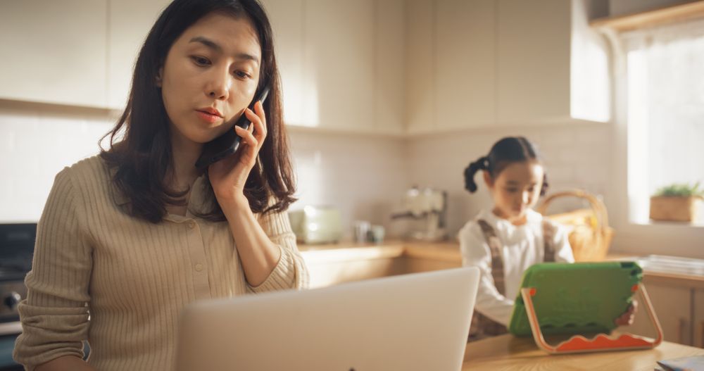 Portrait of Korean Mother Using Laptop and Smartphone To Work from Home While her Little Daughter is Using Digital Tablet.