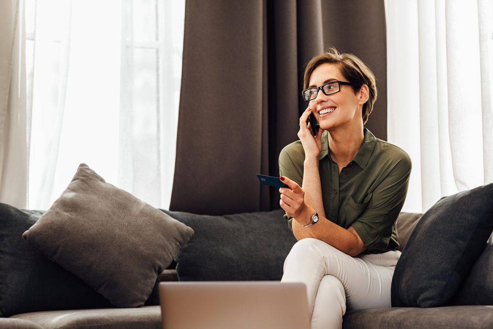 Smiling woman on sofa with pillows with cell phone, credit card and laptop