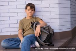 Boy sitting on ground in school corridor next to backpack 5nADl0