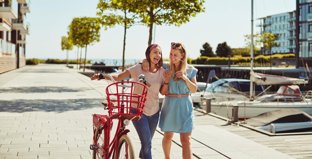 Two happy female friends laughing while walking with bike next to the river