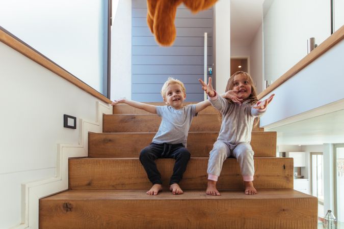 Smiling little kids sitting on stairs and outstretching their hands to catch a teddy bear