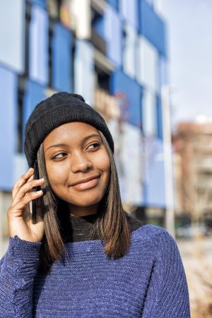 Female in wool hat and blue sweater chatting on cell phone and looking to her side, vertical