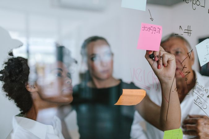 Business woman writing on sticky note on glass wall with coworkers standing by in office