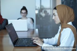 Woman wearing hijab working on her laptop in office with two other women 4NrBr4