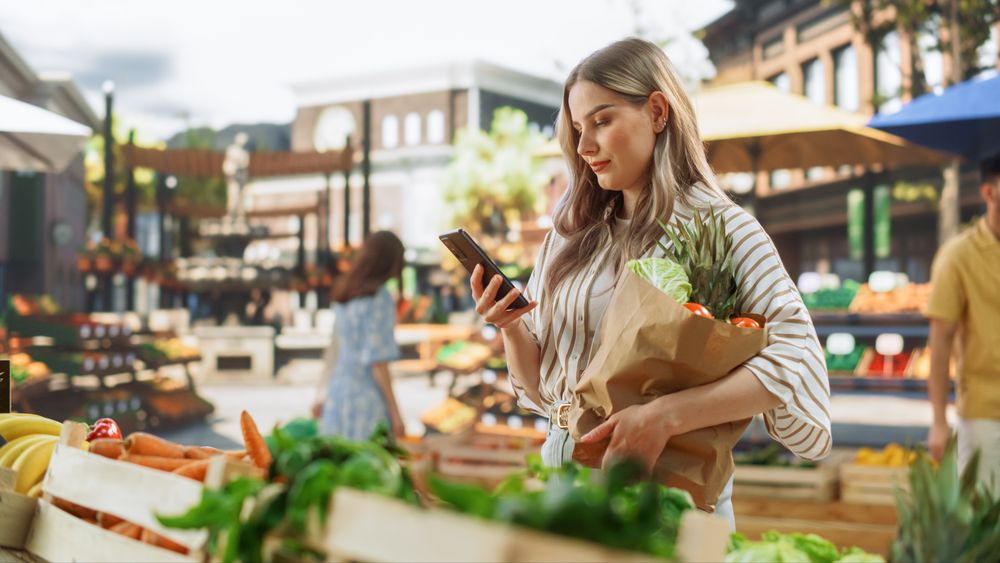 Young Customer Shopping for Fresh Seasonal Fruits and Vegetables, Using Smartphone to Browse Internet on the Move.