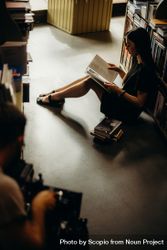Side view of young woman sitting between bookshelves reading a book 5zpjNb