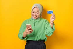Muslim woman in headscarf and green blouse holding credit card and looking down at her smart phone