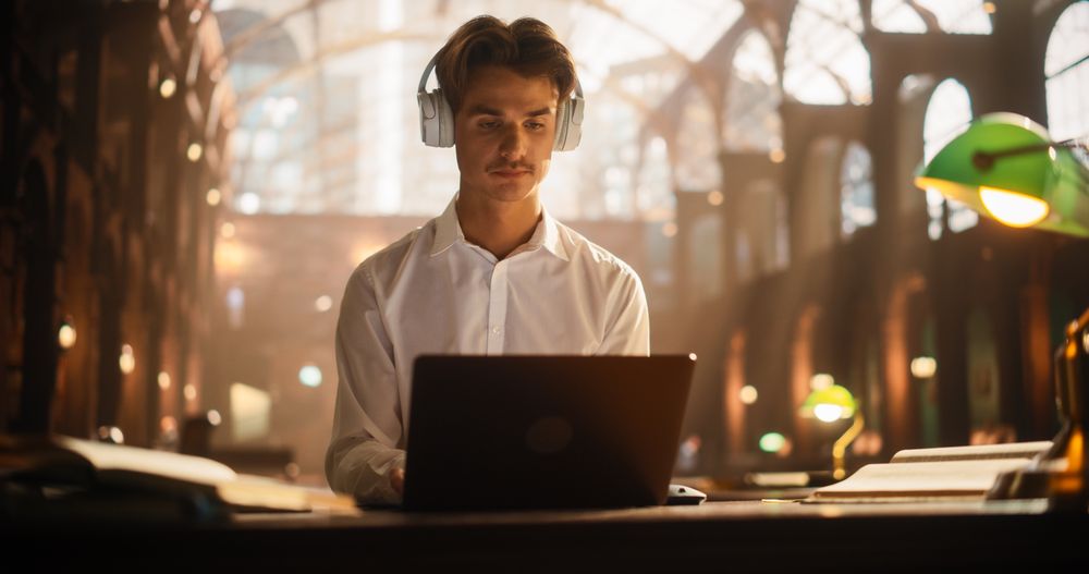 Concentrated Young Male Student Engaged in E-Learning With Headphones and Laptop in a Historic Library