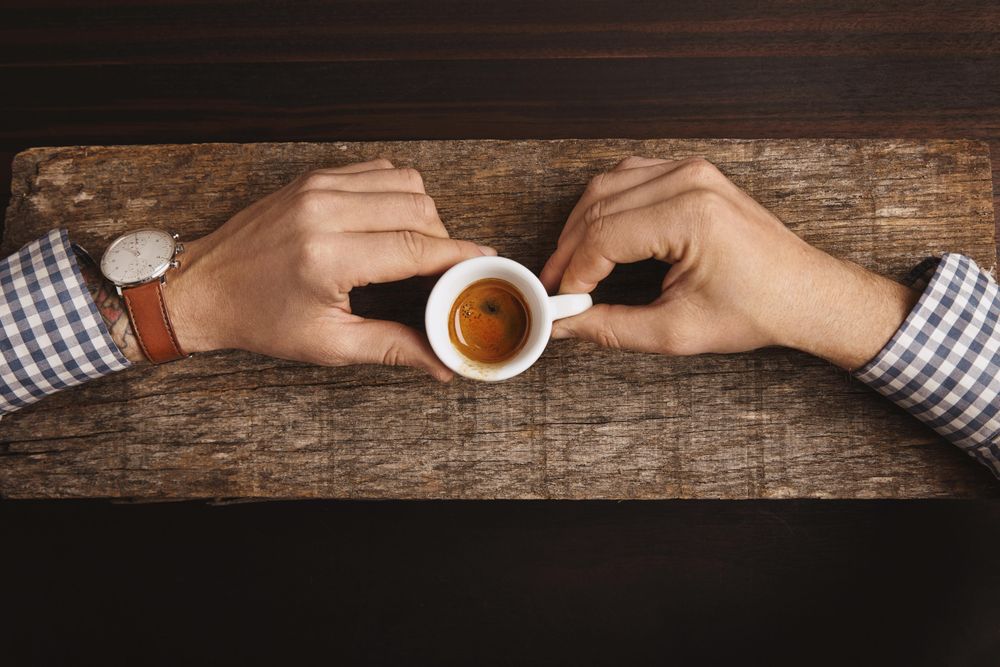 Man wearing plaid shirt and watch holding espresso shot on wooden board