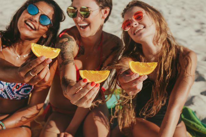 Women in bikinis sitting on beach posing for photograph holding pieces of orange