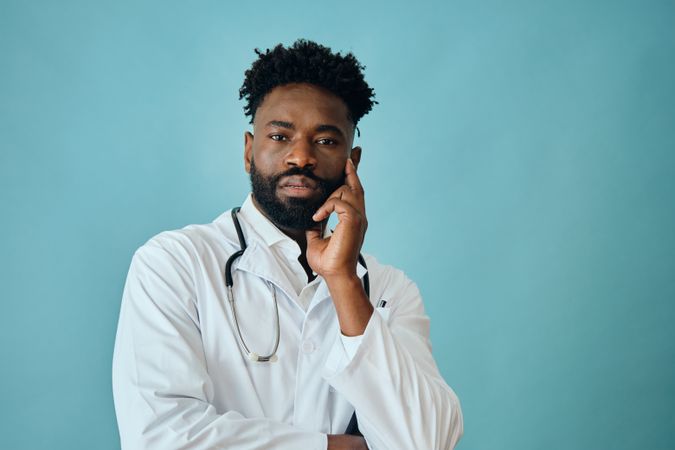 Thoughtful Black male doctor in blue studio with hand to his cheek