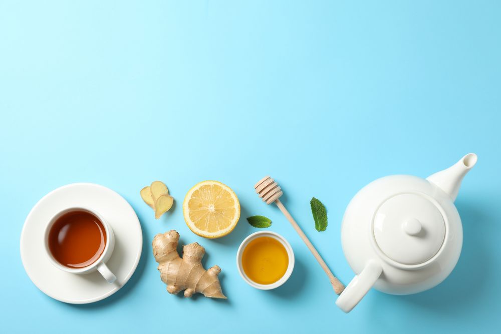 Flat lay. Cup of tea, teapot, lemon, ginger, mint, honey and dipper on blue background, copy space