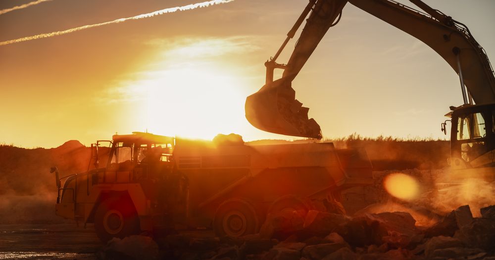 Construction Site On Sunny Evening: Industrial Excavator Loading Rocks Into A Truck. The Process Of Building Apartment Complex. Workers Operating Heavy Machinery To Complete A Real Estate Project.