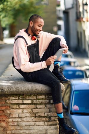 Male sitting with one leg up on brick wall in city reading tablet