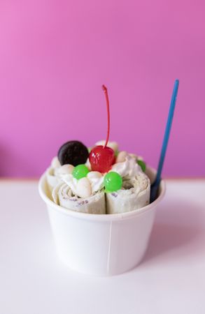 Close up of a rolled ice cream decorated with a cherry, a cookie and green bubble toppings