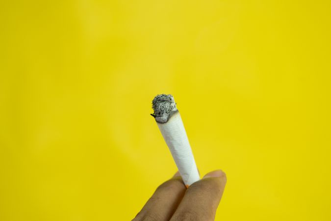 Fingers holding lit hand rolled cigarette on yellow background