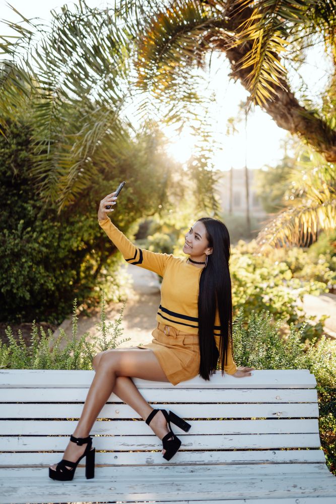 Charming long haired Asian woman with smartphone sitting on bench in park