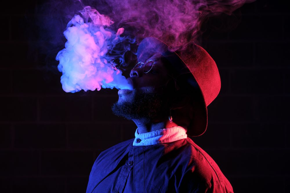 Stylish Black man in hat vaping against brick wall