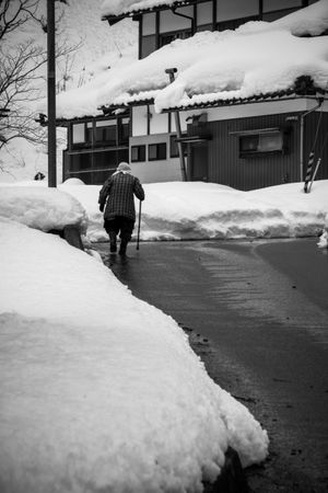 Back view of older person walking in snow-covered neighborhood in grayscale