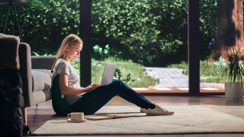 Portrait of Beautiful Young Adult Female with Blond Hair Sitting on a Floor and Using Laptop Computer in Sunny Living Room.