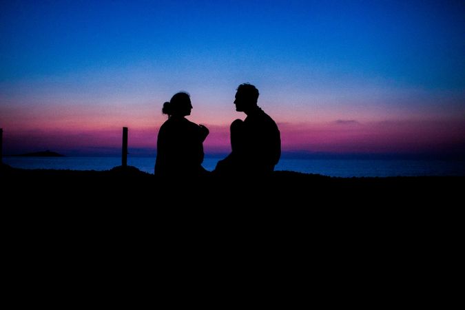 Silhouette of man and woman sitting on seashore at sunset