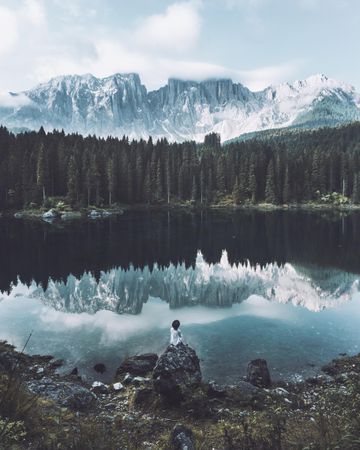 Person sitting on rock near Lake Carezza and green trees and snow covered mountains in Dolomites, South Tyrol, Italy