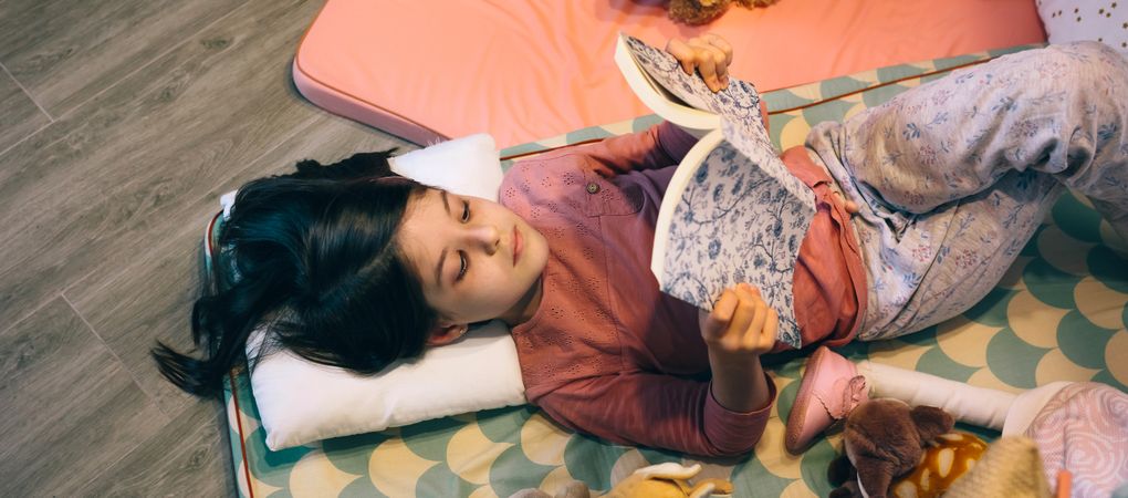 Banner of erene little girl looking a book while resting over cozy sleeping mats in living room