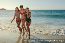 Three girlfriends in bikinis wearing sunglasses walking on beach holding each other bxynv0