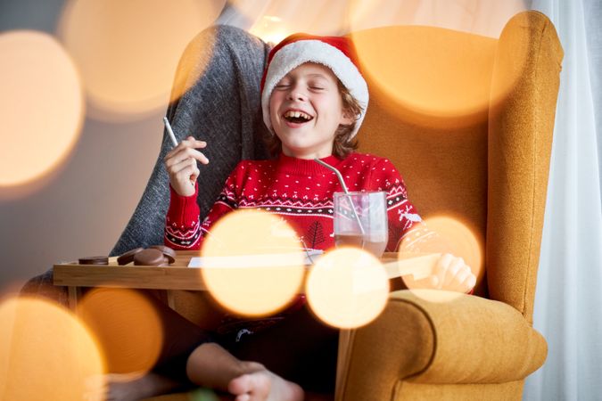 Merry kid painting in armchair and enjoying chocolate during Chr