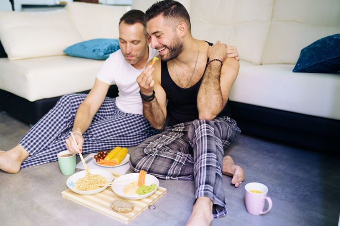 Happy male couple enjoying relaxing meal at home sitting on floor