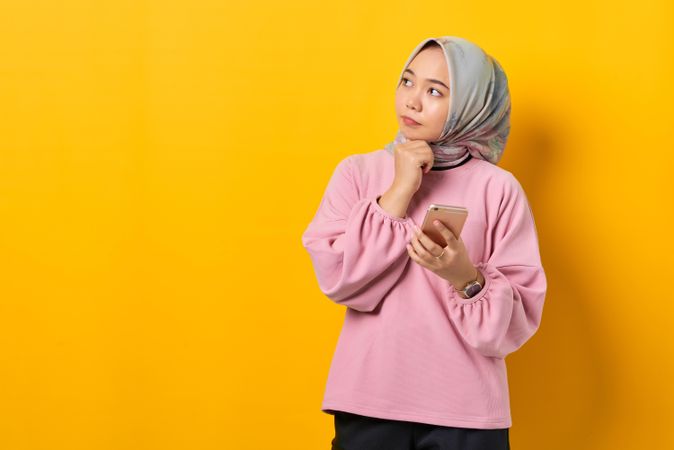 Muslim woman in headscarf holding smart phone and looking away in thought with hand on her chin