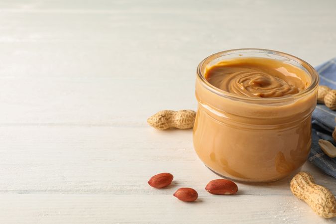 Glass jar with peanut butter, peanut and kitchen towel on wooden background, closeup and space for text