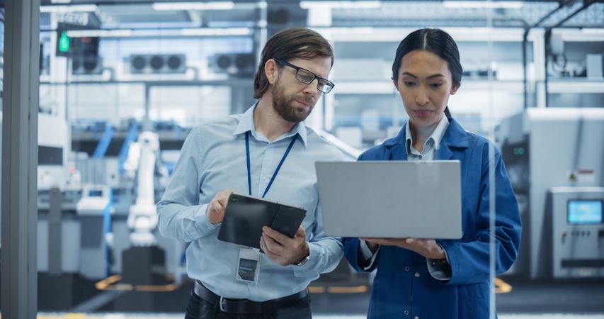 Engineer and Asian Female Project Manager Standing in Office, Talking and Using Computers and Monitoring Production at Modern Automated Electronics Manufacture with AI Robotic Arms