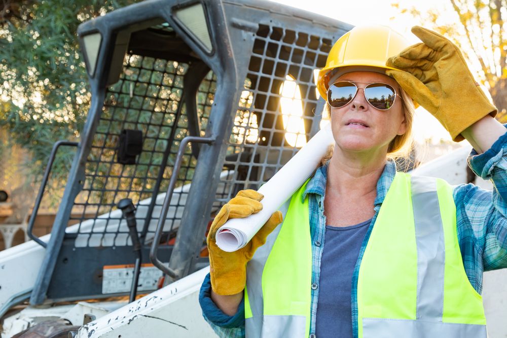Female Worker Holding Technical Blueprints Near Small Bulldozer At Construction Site