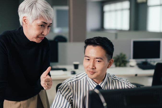 Mature Asian woman reviewing project on computer with male colleague and giving thumbs up
