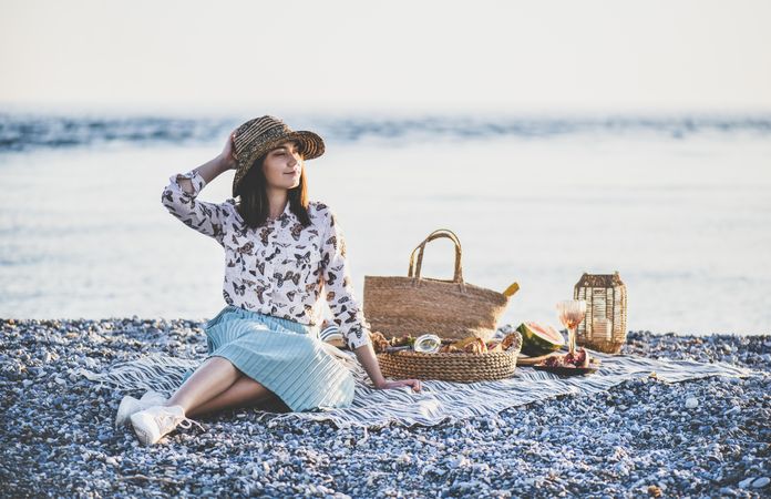Young woman in hat sitting by the ocean with a picnic, watermelon, and wine looking away