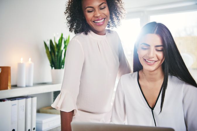 Two female colleagues in a bright office smiling looking at a laptop