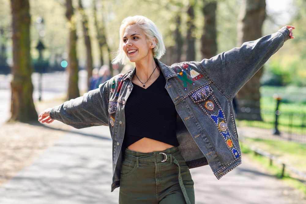 Blonde woman in jean jacket with arms outstretched while walking in park