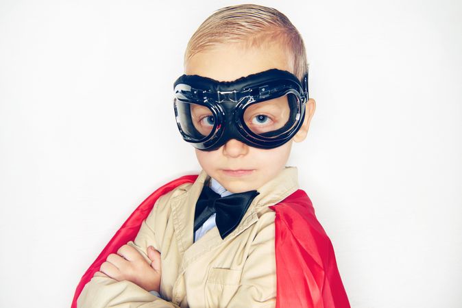 Serious blond boy wearing airplane goggles and cape with arms crossed