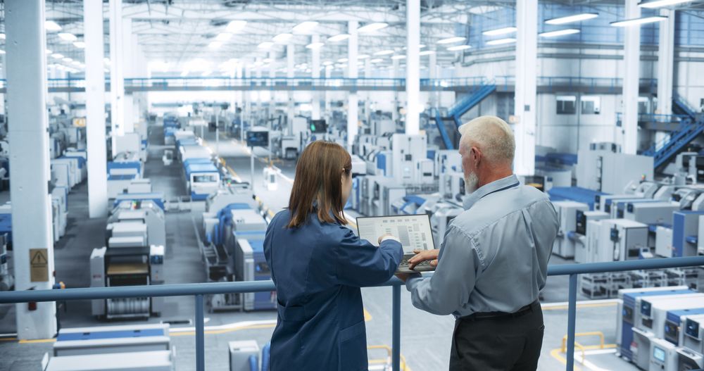 Portrait of a Mature Engineer and Happy Asian Scientist Using Laptop Computer and Talking in a Factory Facility with Equipment Producing Modern Electronic Components for Different Industries