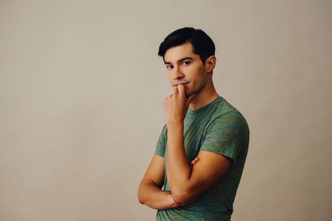 Contemplative Hispanic male facing left in neutral room with arms crossed and finger over mouth