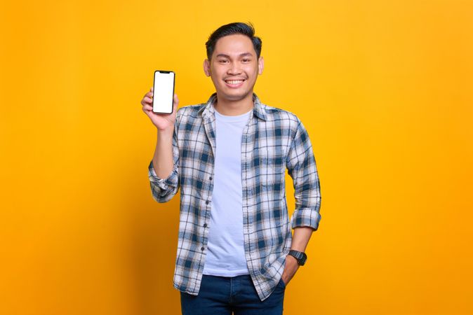 Smiling Asian male with hand in pocket and showing blank screen of smart phone