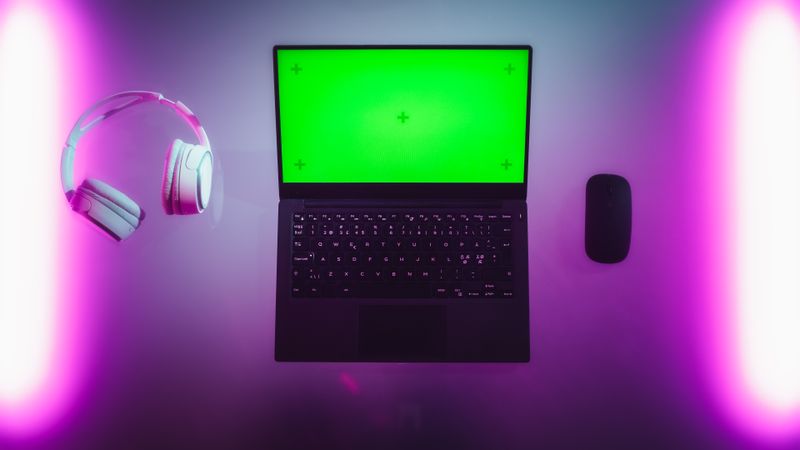 Top Down View of a Laptop Computer with Mock Up Green Screen Chromakey Display with Motion Trackers Standing on an Neon Desk Next to Wireless Mouse and Headphones. Online Gaming Concept