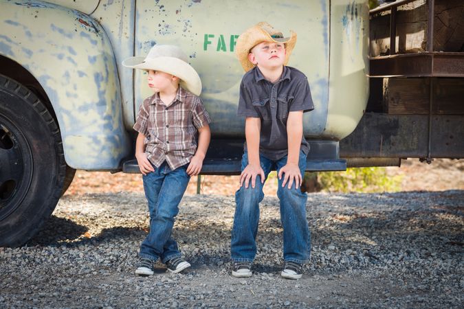 Two Boys Wearing Cowboy Hats Leaning Against an Antique Truck in a Rustic Setting