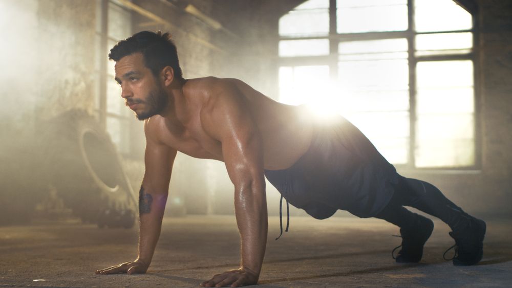 Muscular Shirtless Man Covered in Sweat Does Push-ups in a Deserted Factory Remodeled into Gym.