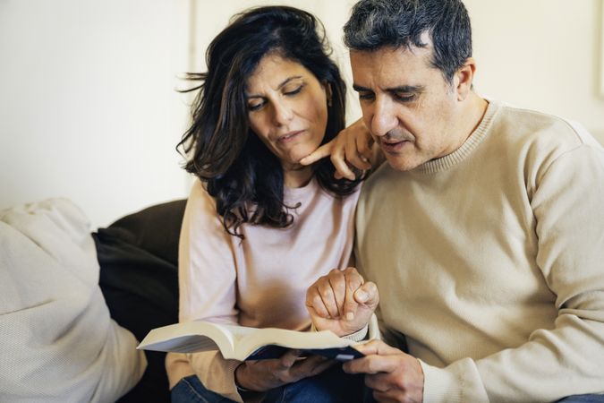 couple relaxing on sofa reading a book together and pointing at text with curiosity