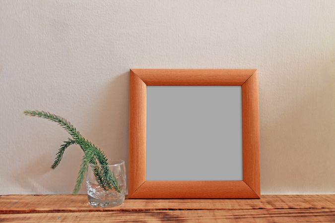 Plain square wooden picture frame with grey interior leaning against wall with plant in glass mockup